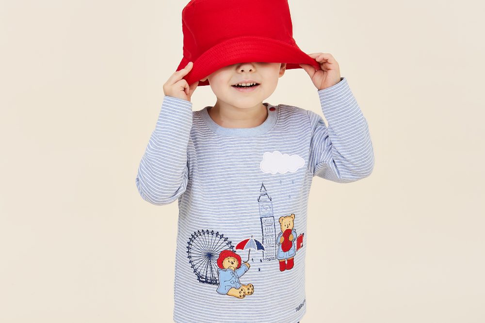 A child pulling a red hat over their eyes wearing a top with Padding Bear on from the JoJo x Paddington collection