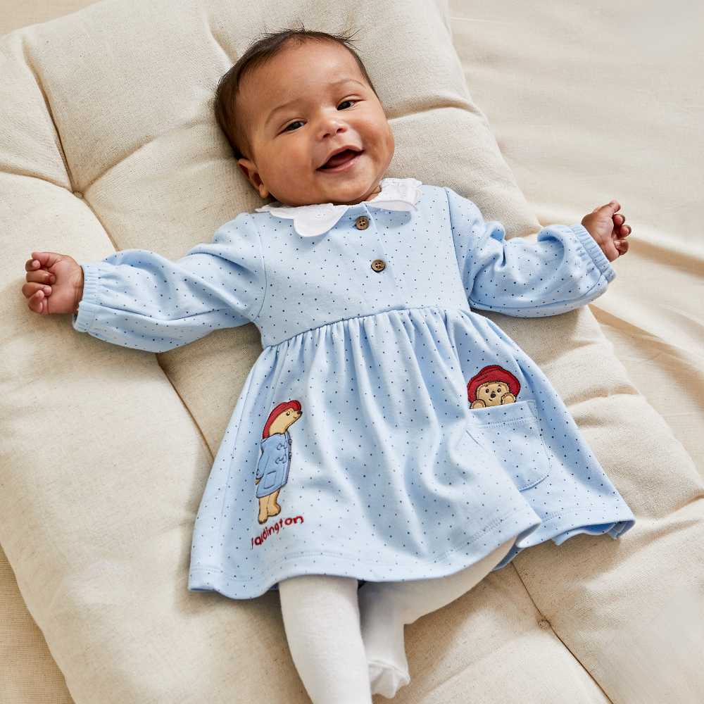 A baby lying down wearing a blue dress with Padding Bear designs on the front 