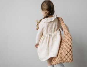 The back of a girl in a cream dress with long hair in a plait and a string bag over her shoulder