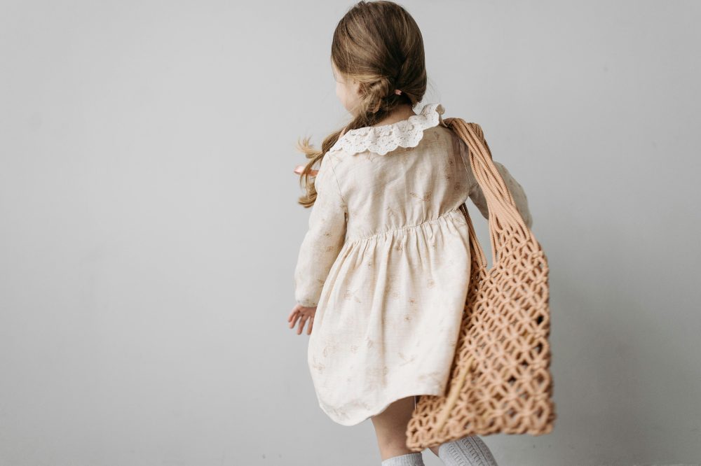 The back of a girl in a cream dress with long hair in a plait and a string bag over her shoulder