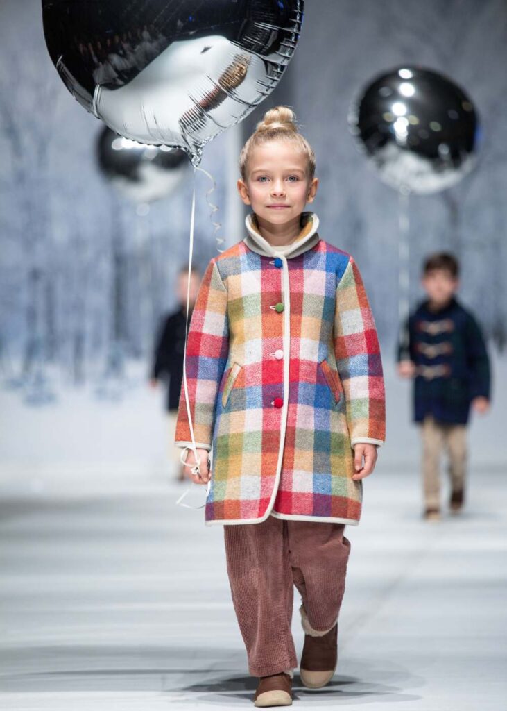 A child stood on a fashion catwalk in a checked coat holding a silver balloon