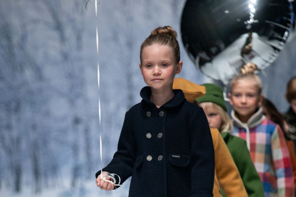 A row of children walking down a fashion catwalk holding silver balloons