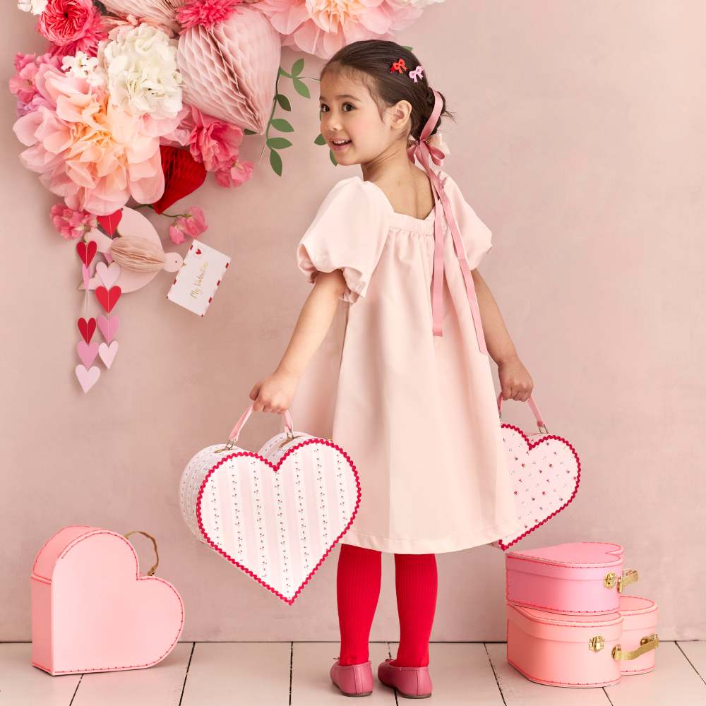 A girl in a pink dress stood beside a large paper flower display with her back to the camera holding heart-shaped gift cases