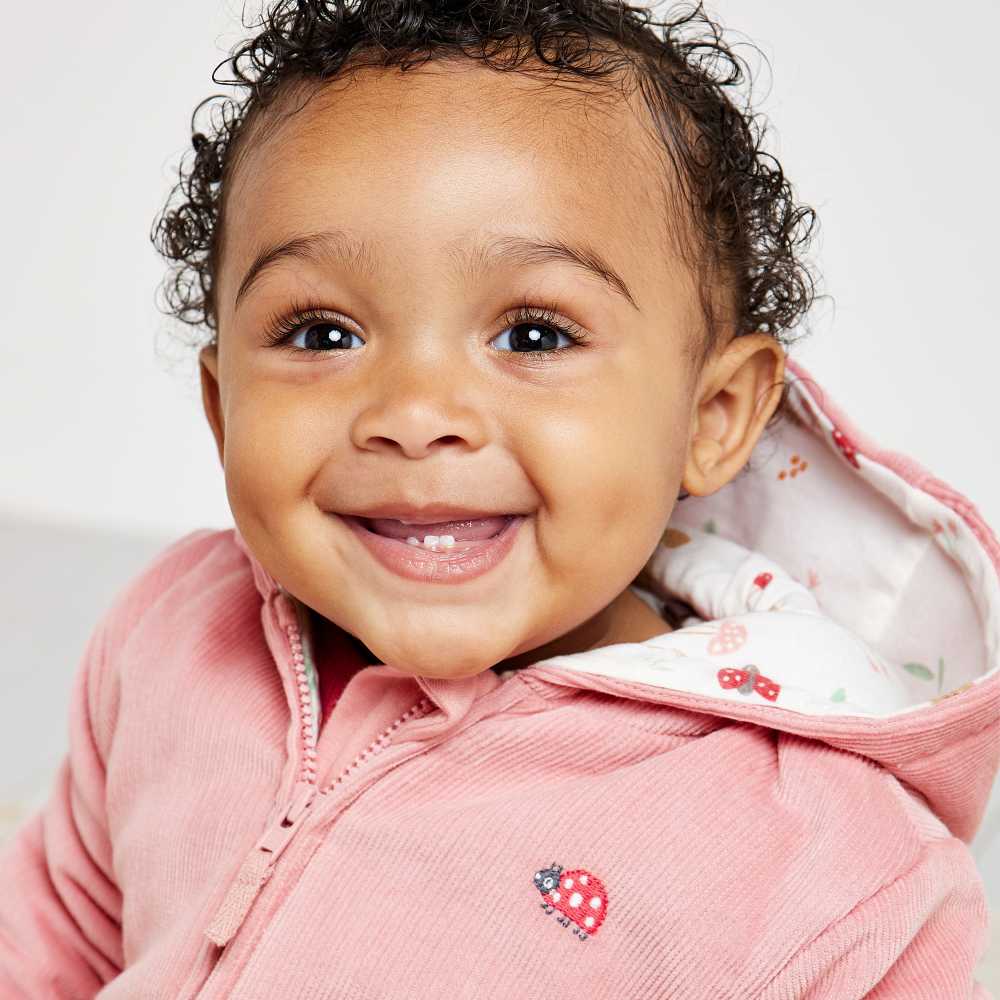 A baby smiling at the camera wearing a pink hooded jacket with a ladybird on 