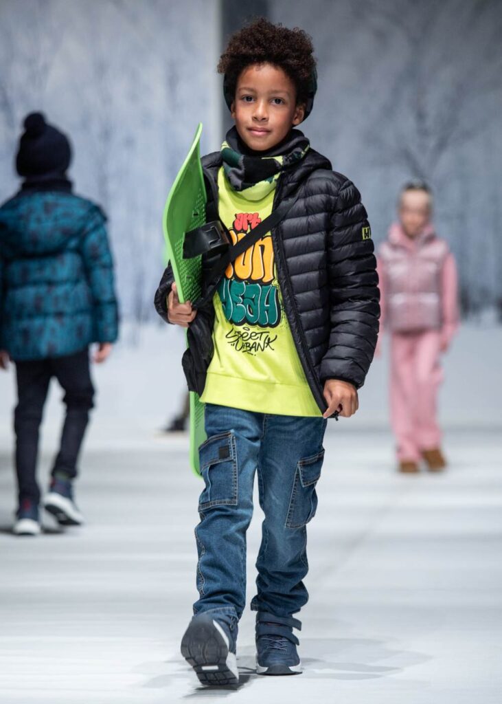 A boy in a black padded jacket, yellow T-shirt and jeans walking down a catwalk holding a green skateboard 