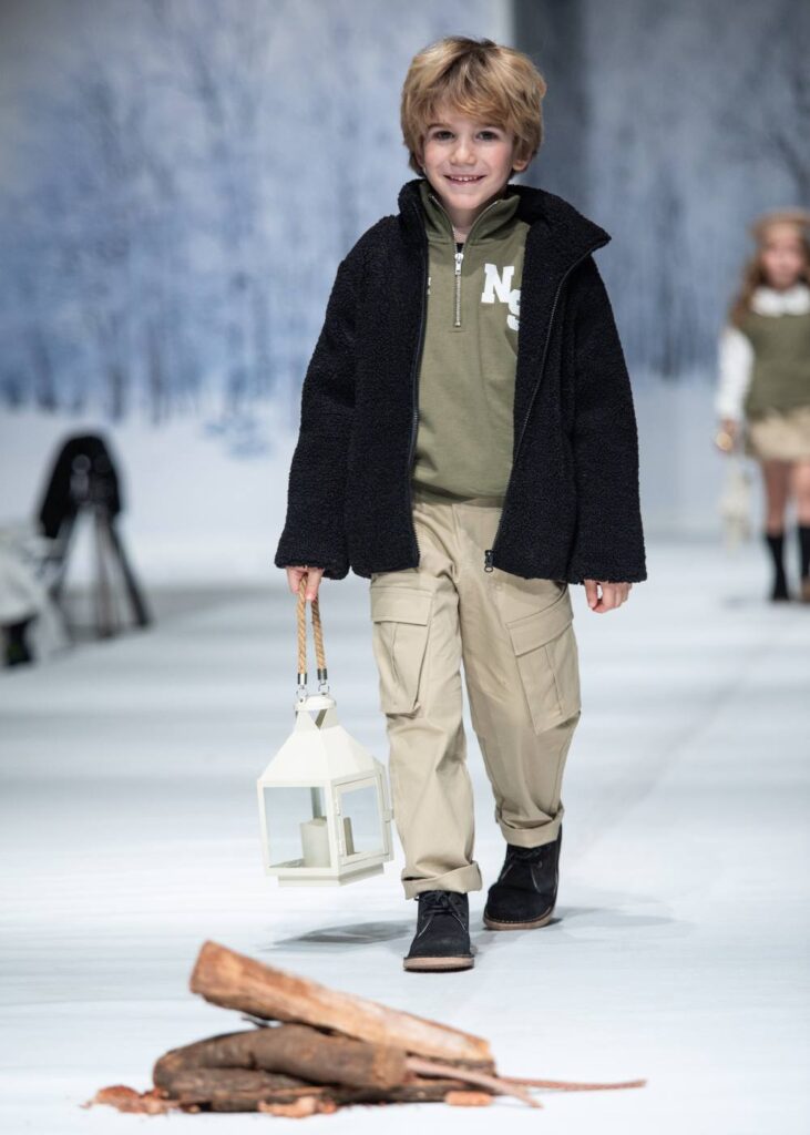 A boy in a dark coat, green top and beige trousers walking down a catwalk holding a lantern 