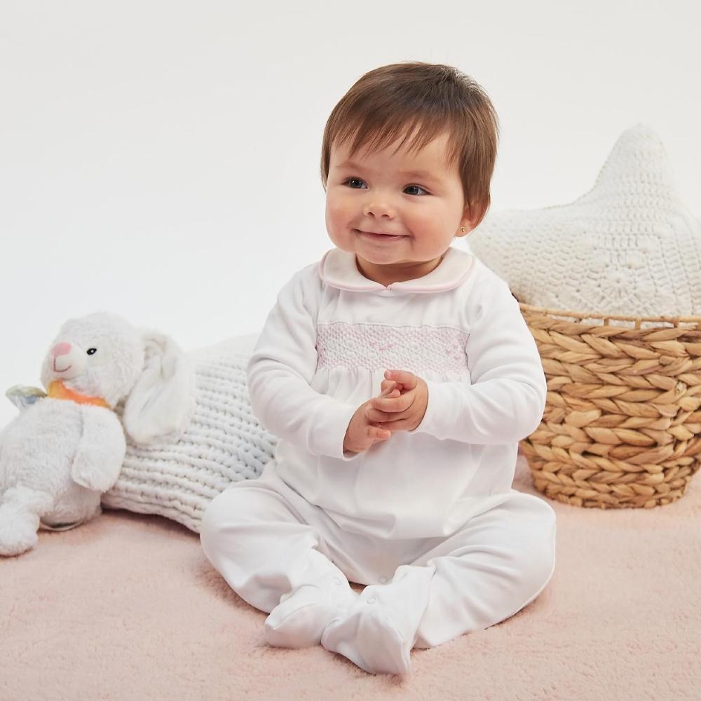 A young baby in a white outfit sat on the floor beside a wicker backet and a soft toy 