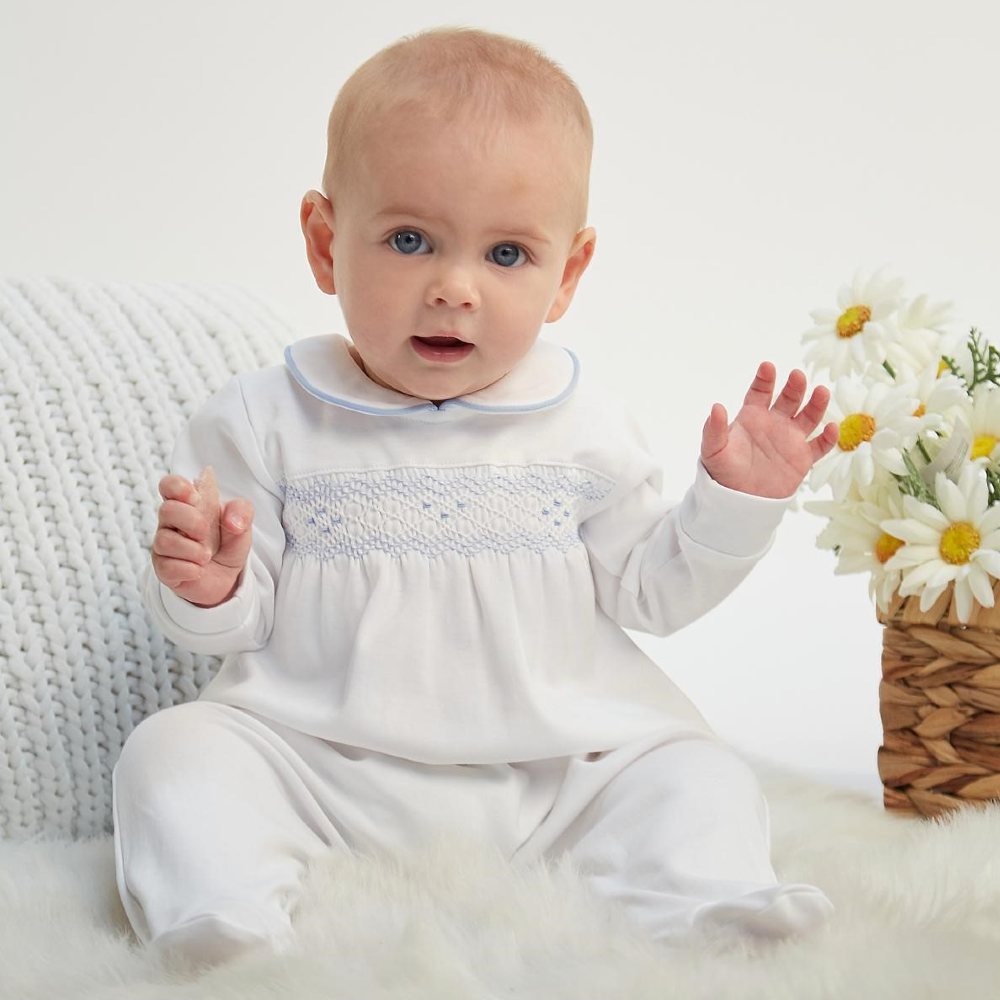 A baby wearing a white outfit sat beside a knitted cushion and a vase of daisies 