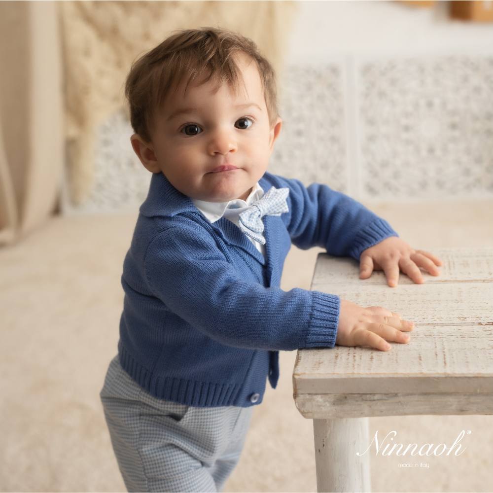 A toddler stood holding onto a table wearing a blue knitted cardigan and blue trousers 