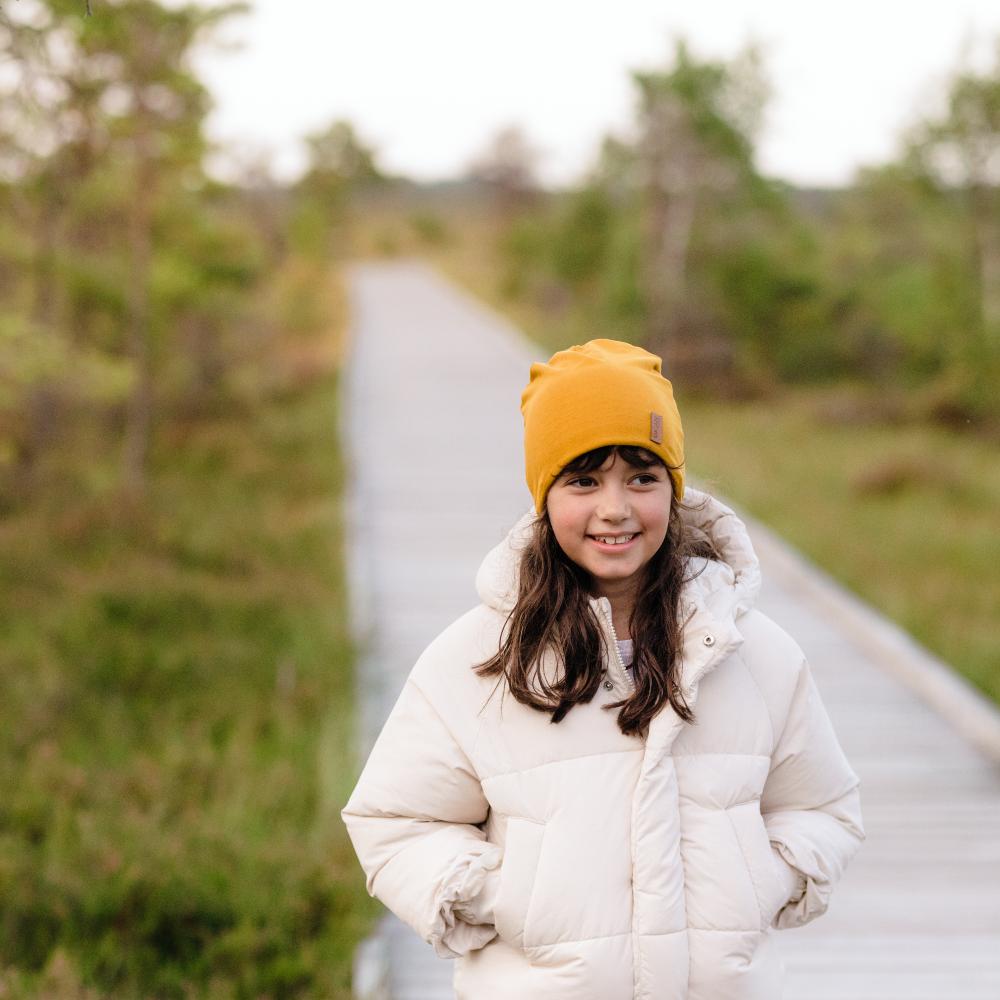 A girl stood outside on a pathway wearing a cream padded jacket and yellow beanie hat 