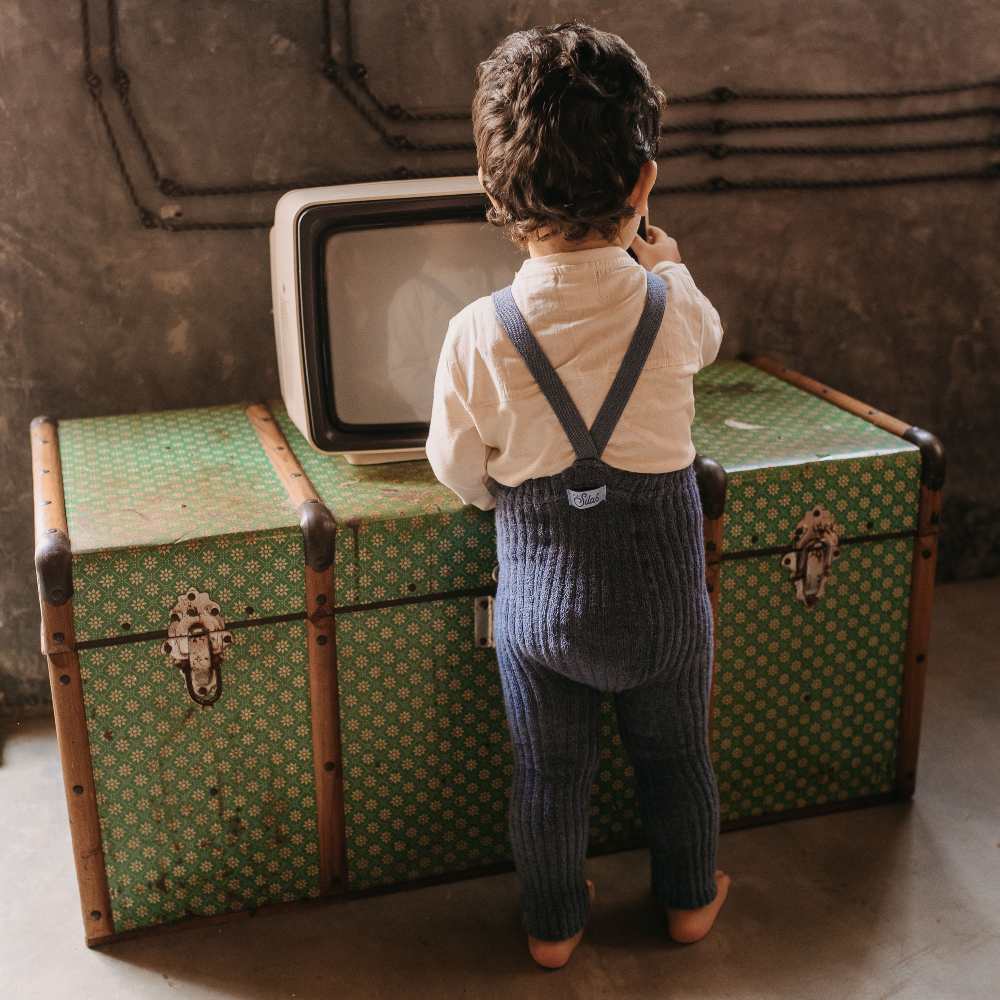 A young child stood in front of a green chest with a TV on wearing blue knitted leggings with braces 