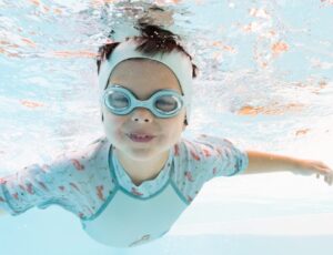 A child underwater in a pool wearing swimming goggles and a ear band by Splash About