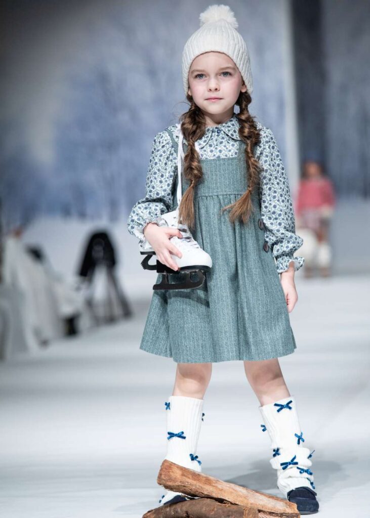 A girl on a catwalk holding ice skates wearing a white bobble hat , blue dress and white knee socks with blue bows on 