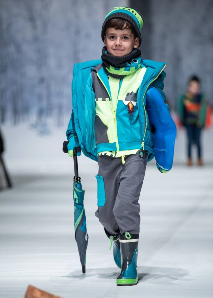 A boy in a blue hat, jacket and jumper with grey trousers walking down a catwalk holding a blue umbrella 