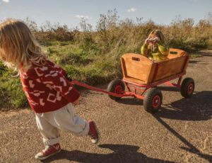 A child in a red jumper pulling a wooden cart with a child in