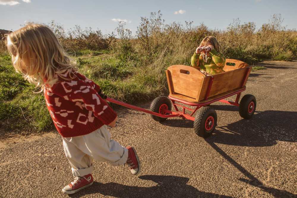 A child in a red jumper pulling a wooden cart with a child in