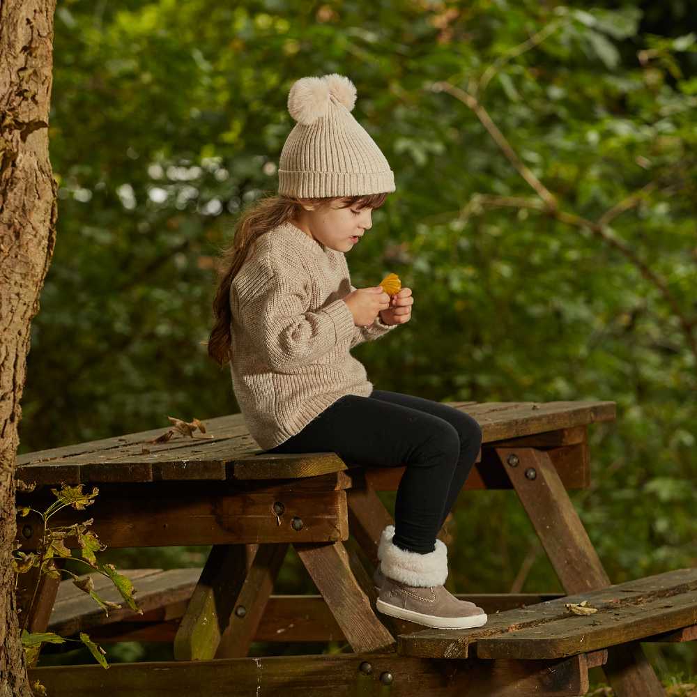 A girl sat outside on a picnic table wearing a bobble hat and winter boots 