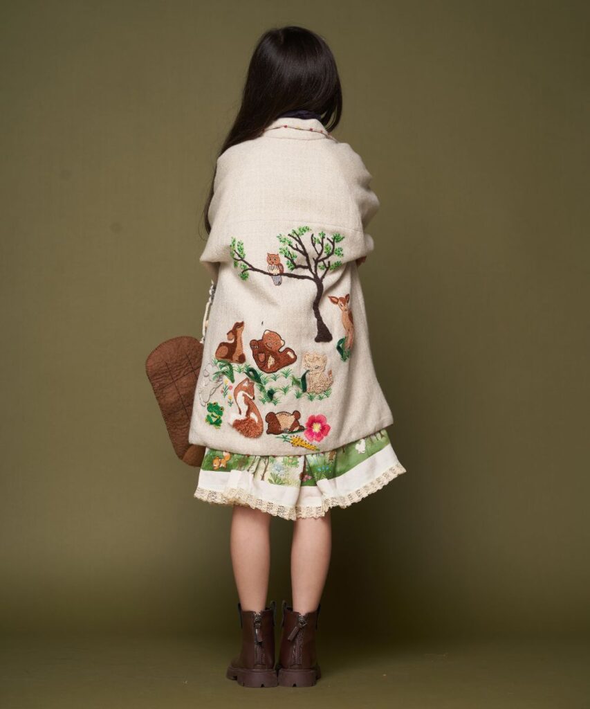 A girl with dark hair with her back to the camera wearing a cream coat and skirt with woodland motif embroidery 