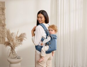 A woman in a room beside carrying a young child on her back in a blue BabyBjorn carrier