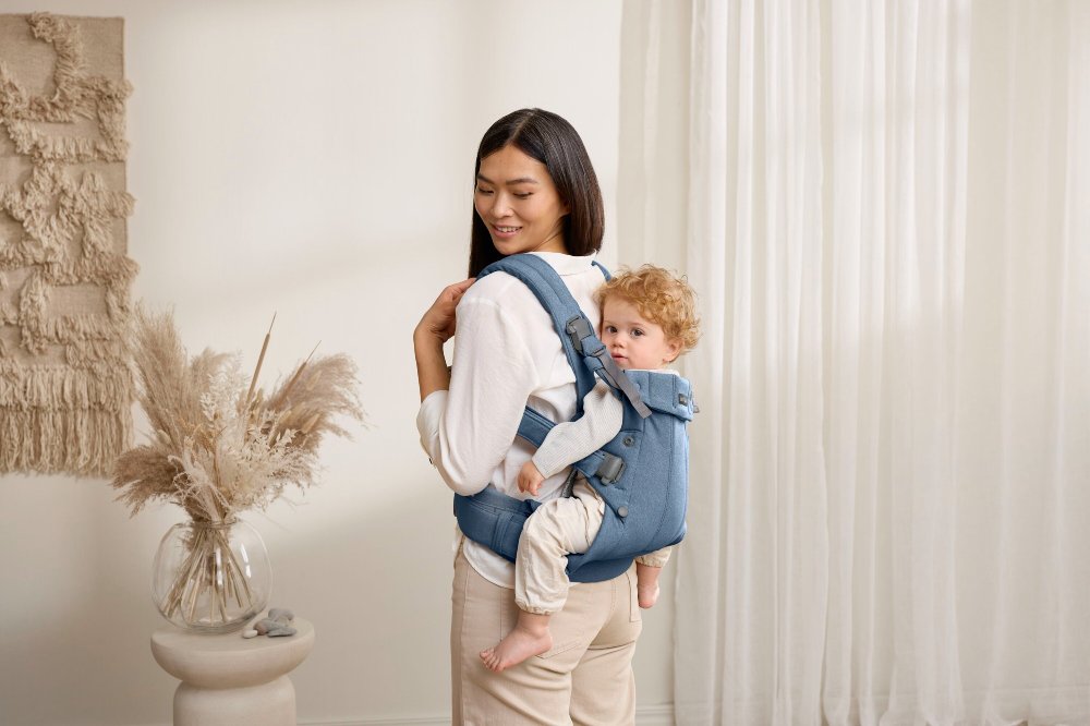 A woman in a room beside carrying a young child on her back in a blue BabyBjorn carrier