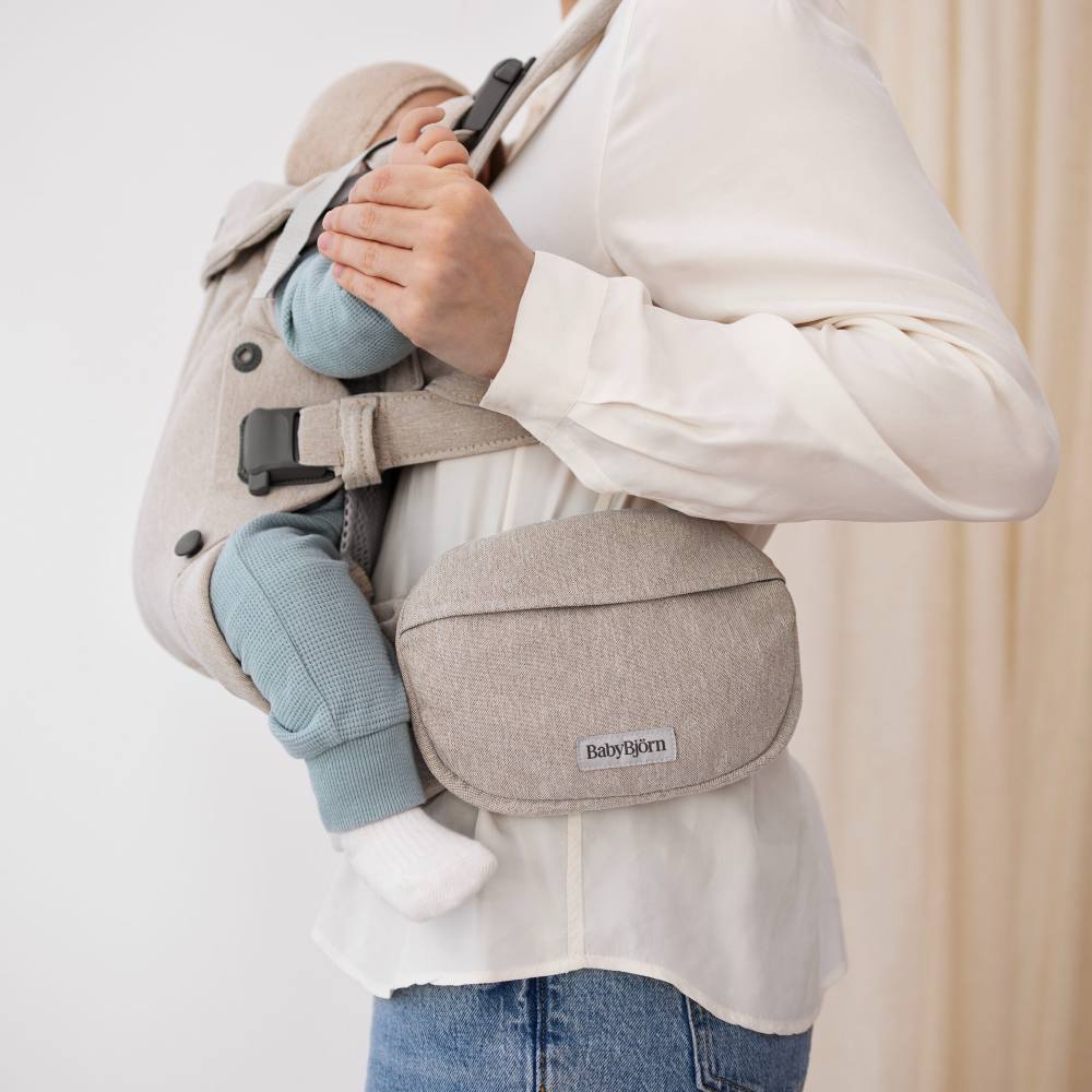 A woman in a white shirt and jeans with a baby carrier on her front with a pocket pouch on the side 