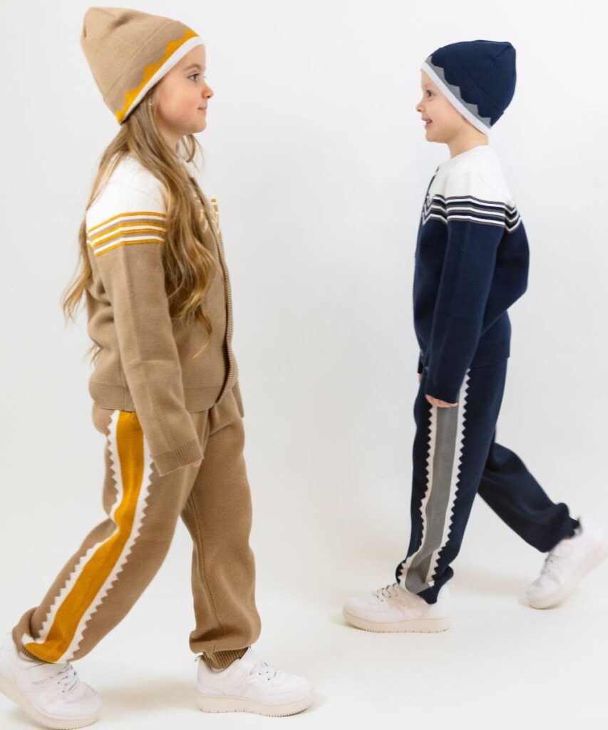 A young girl and boy walking towards each other wearing sports-inspired jackets and trousers and matching beanie hats 