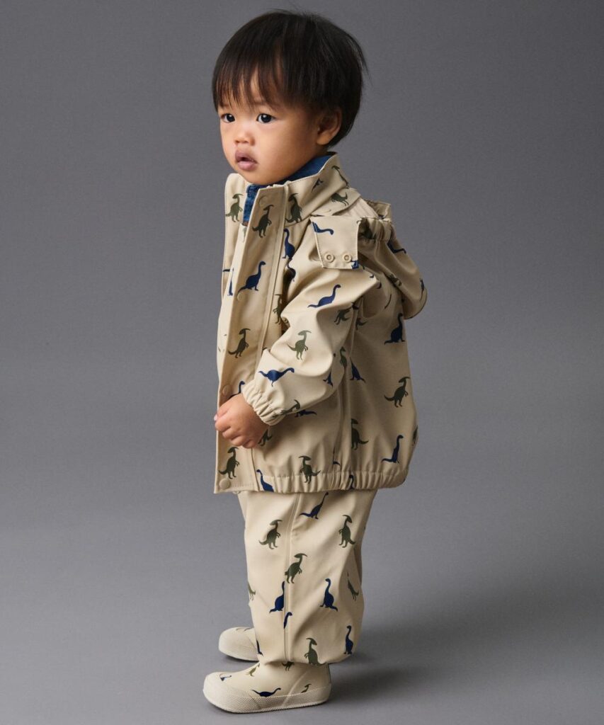 A young child stood against a grey background wearing a matching rain jacket and waterproof trousers 