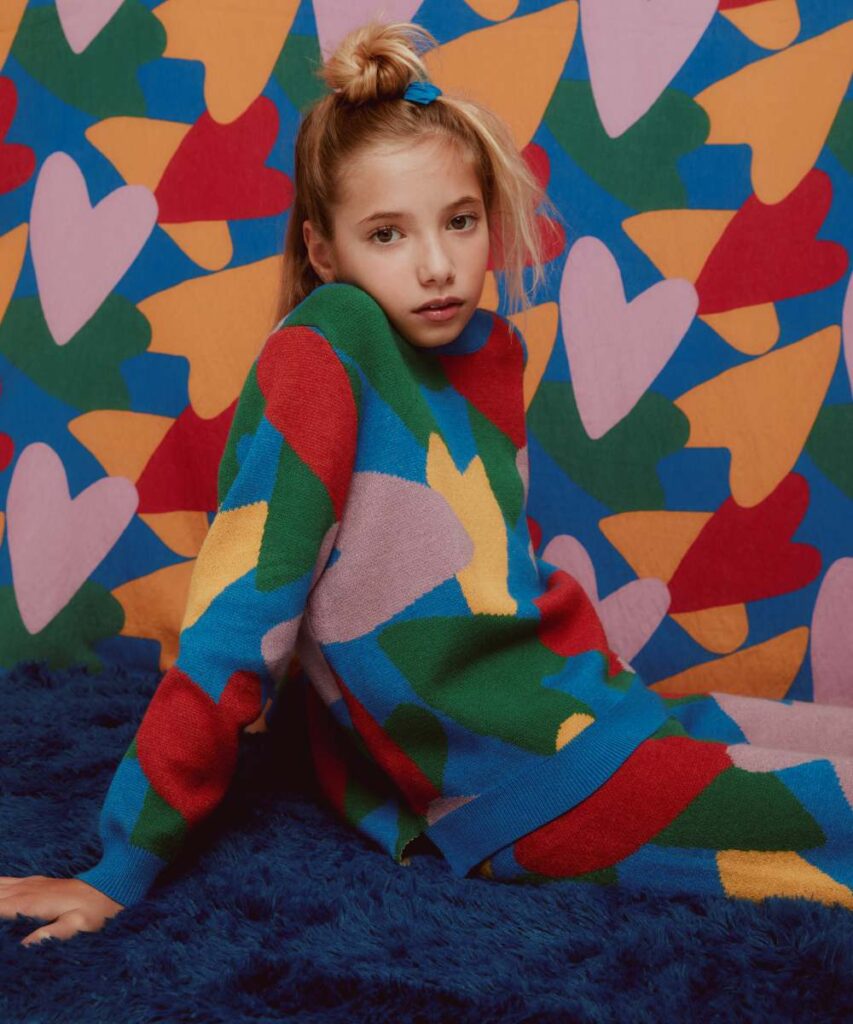 A young girl sat on a blue fur seat wearing a bright heart motif top and trousers with matching heart motif wall paper in the background 