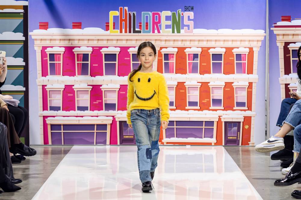 A child in a yellow smiley face jumper and jeans walking down a fashion catwalk at the Children's Show 