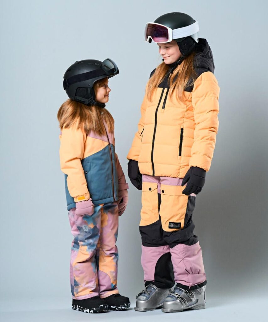 Two young girls stood against a grey background wearing colourful skiwear and helmets and ski goggles on their heads 