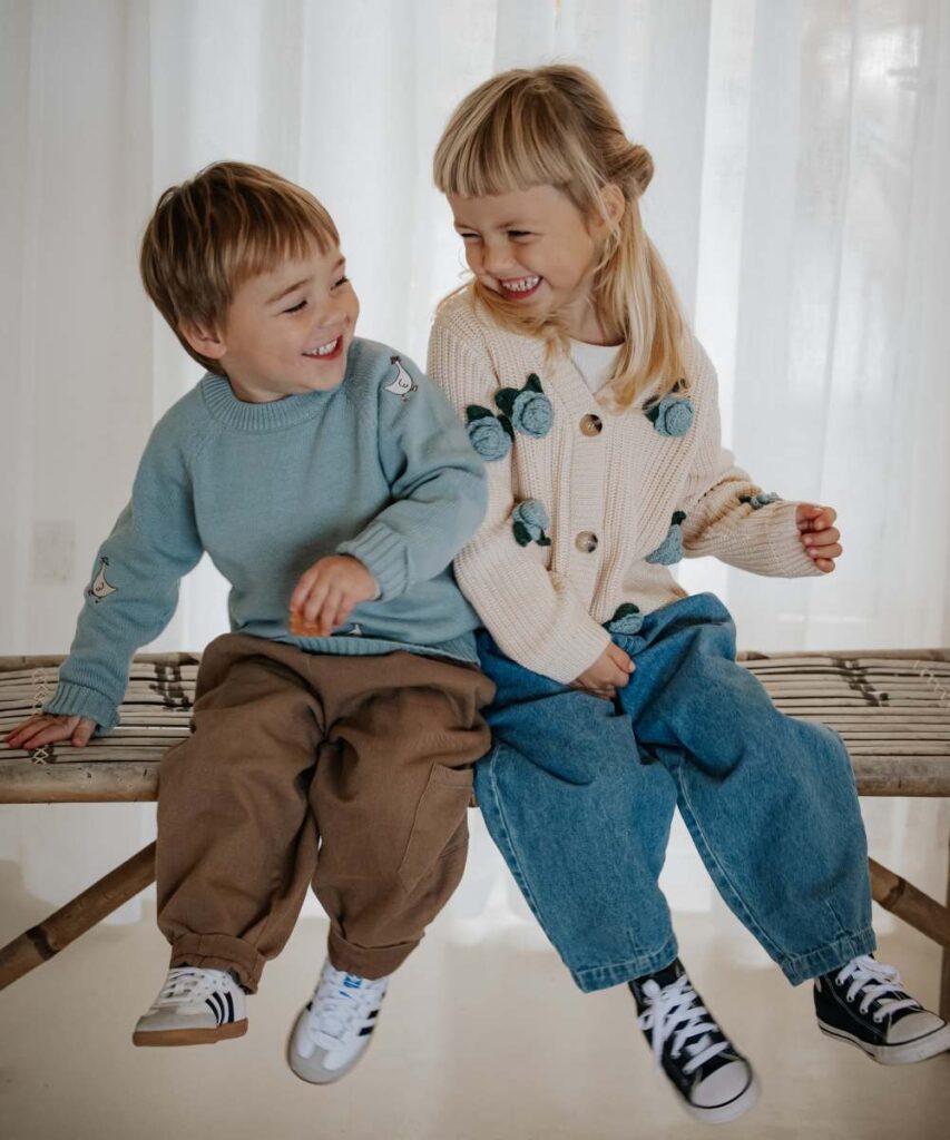 Two young children laughing sat shoulder to shoulder on a wooden bench 