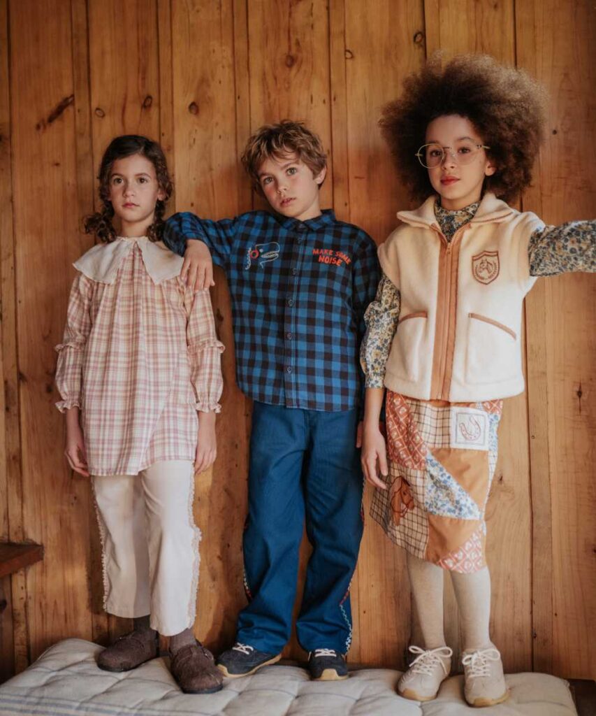 Three children stood side by side on a cushion against a wooden panelled wall 