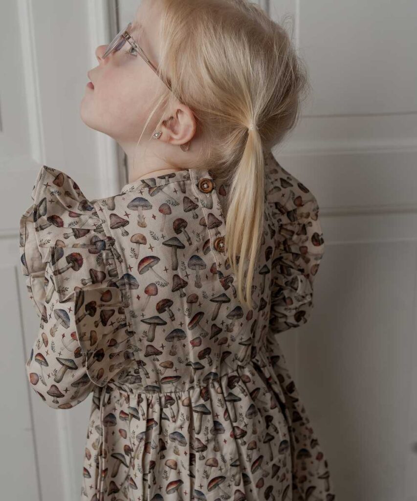 A young girl in glasses with blonde hair in bunches wearing a beige dress with an all-over mushroom print stood against a white wall looking upwards 