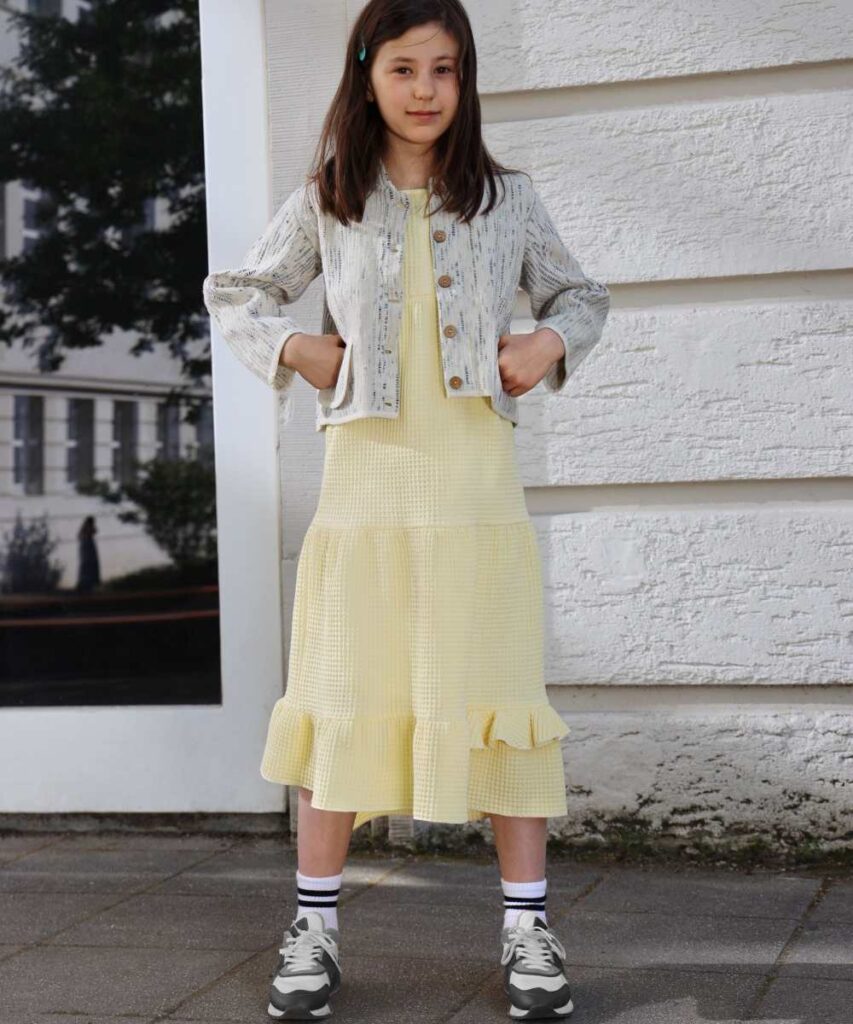 A young girl with dark hair stood outside a building wearing a pale yellow dress, jacket and trainers 