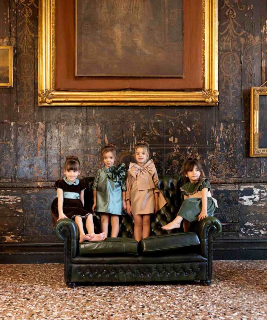 Four young children in partywear stood on a green leather sofa in a grand room 