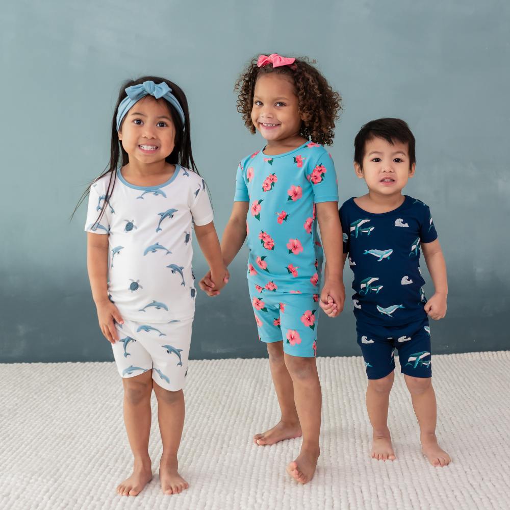 Three children stood in a row holding hands wearing printed shorts and T-shirts by Kyte Baby 