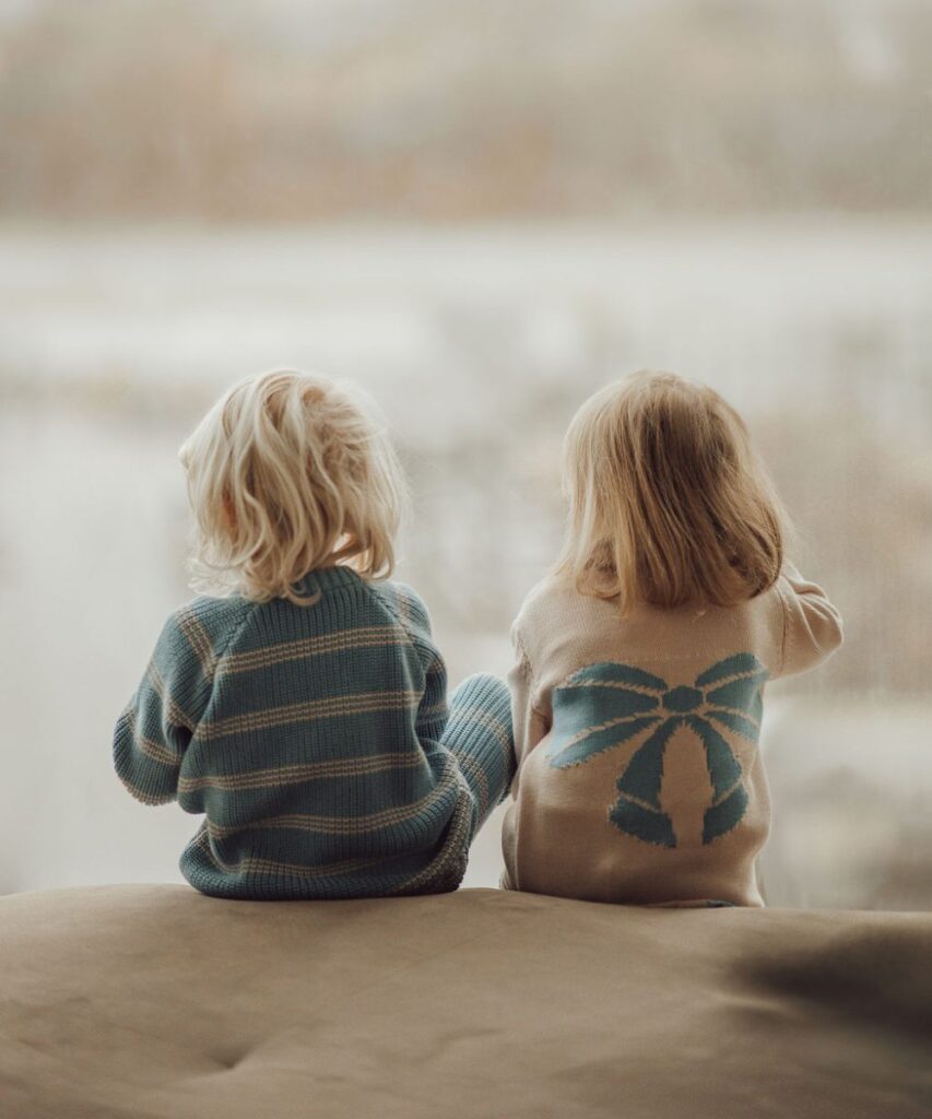 Two young children sat with their backs to the camera wearing a blue striped jumper and a cream jumper with a blue bow design