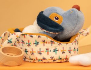 A Lilliputiens dog plush toy in a basket beside a bowl and bone toy