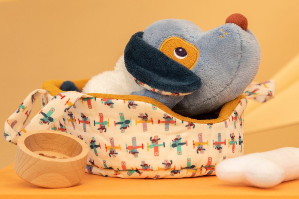 A Lilliputiens dog plush toy in a basket beside a bowl and bone toy