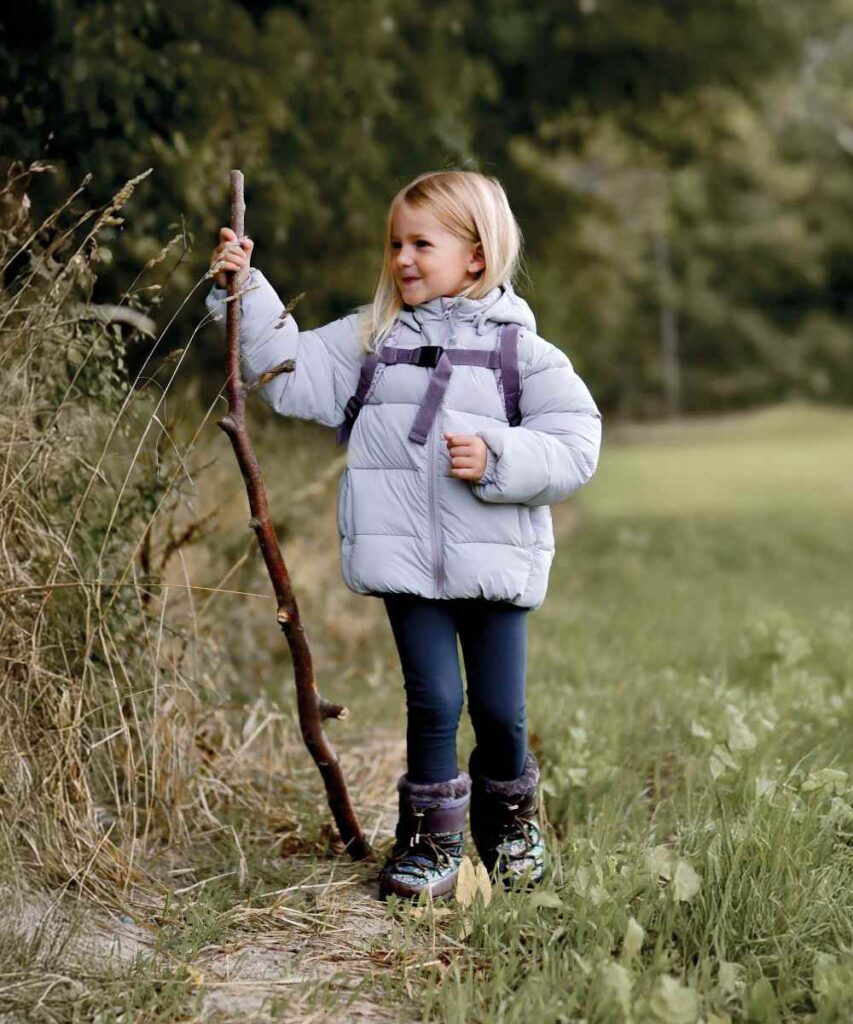 A young girl in a puffer jacket, leggings and winter boots walking in a field holding a large stick 