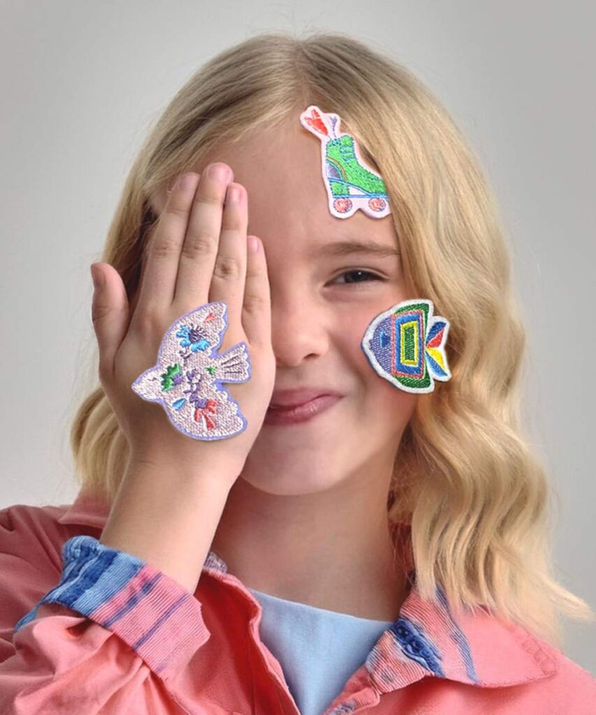 A young girl smiling at the camera with a had over one eye and colourful patches on her hand and face 