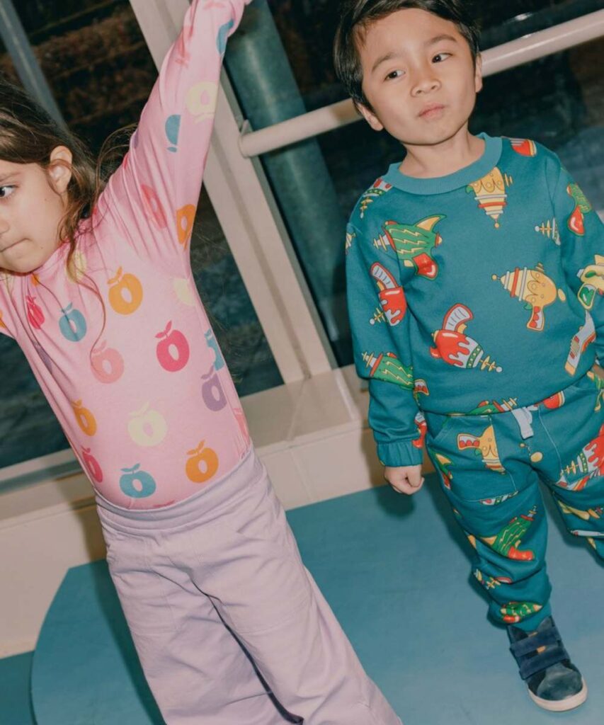 A girl in pink trousers and a pink top with an apple motif print holding her arms up beside a young boy in a blue top and trousers with a space gun print on them