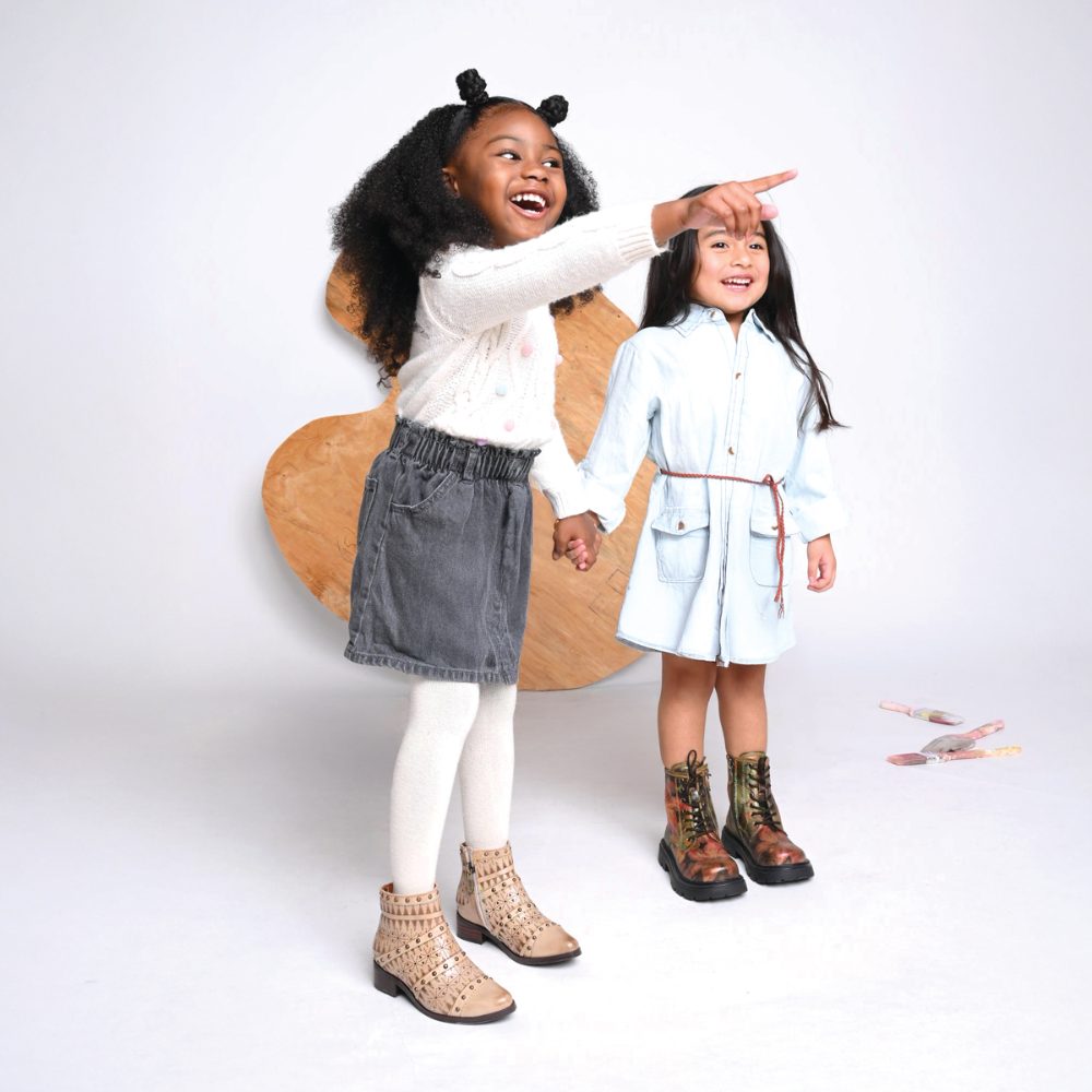 Two girls holding hands with one point their finger and both wearing boots from the L’Artiste Kids Collection 