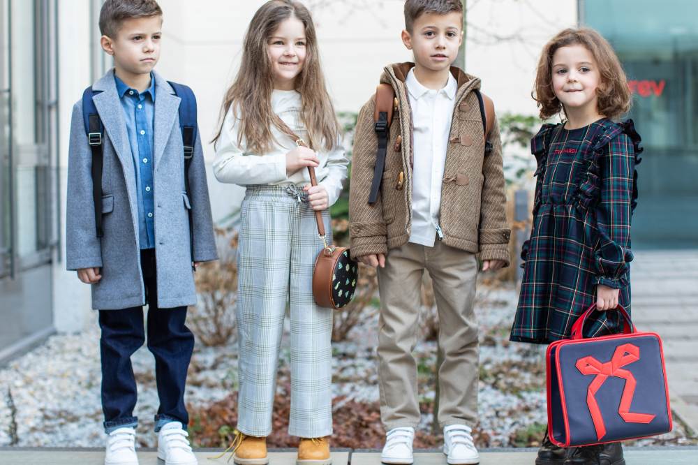 Four children stood outside on a wall with one girl holding a navy bag with a red bow design on the front 