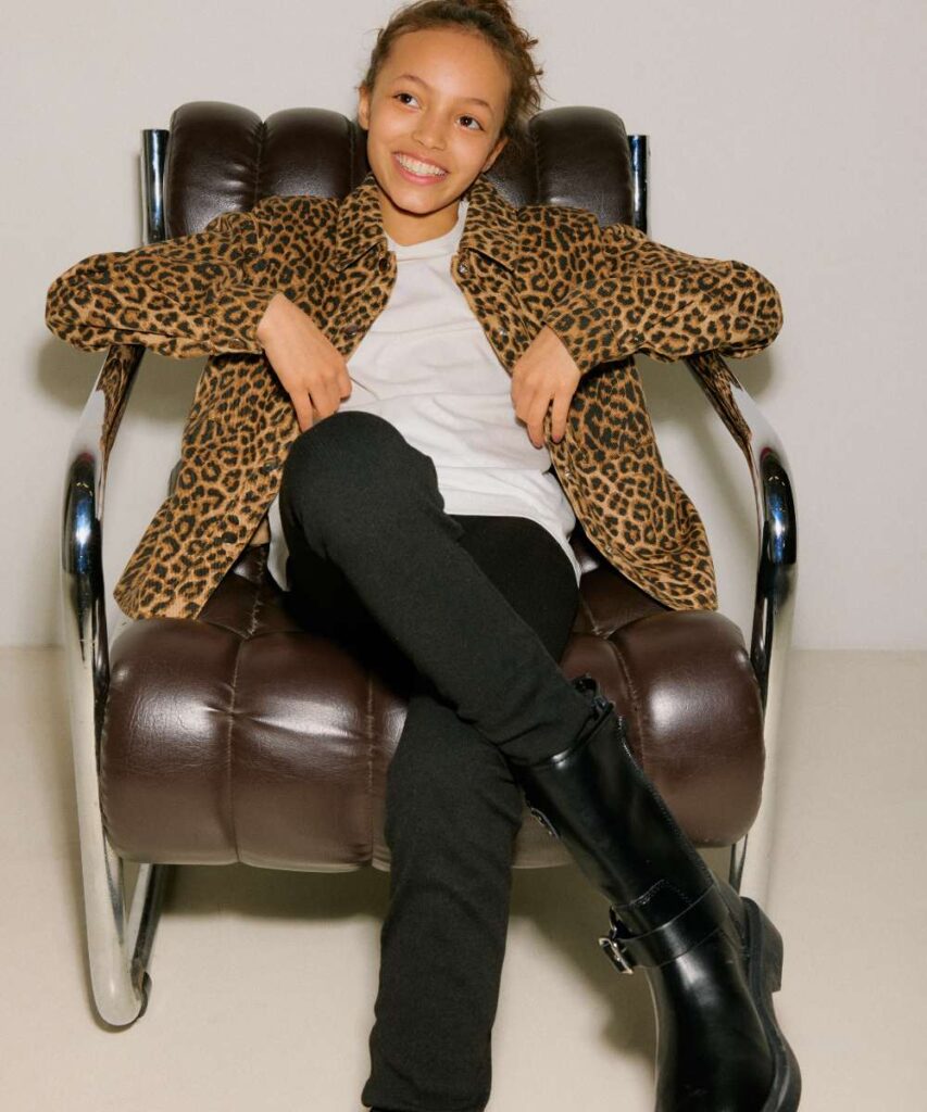 A girls sat in a brown leather chair smiling at the camara wearing a leopard print jacket, white top and black trousers and boots 