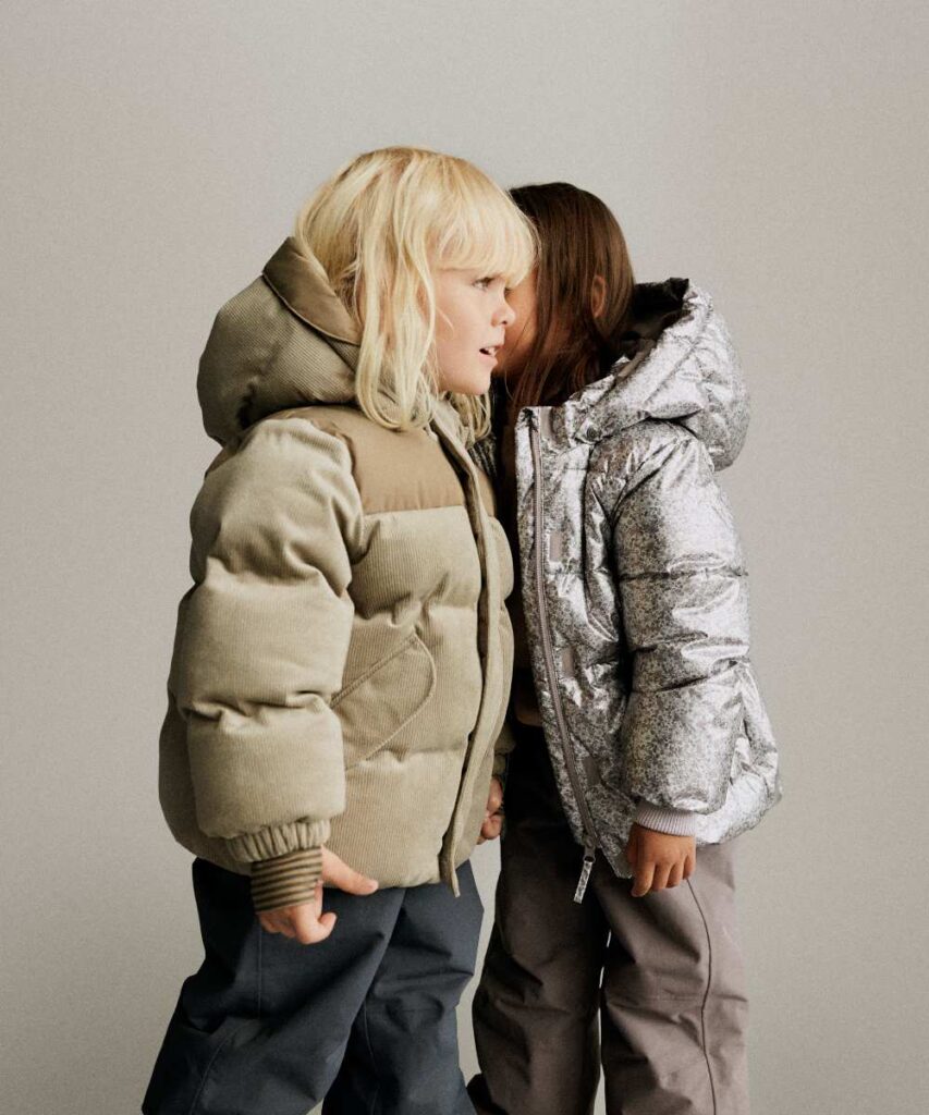 A child in a silver puffer jacket whispering in another child's ear who is wearing a khaki puffer jacket 