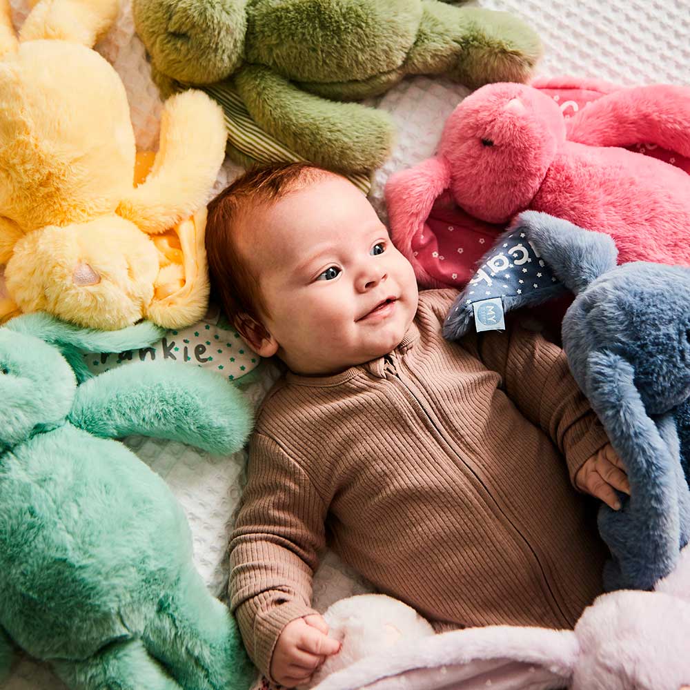 A young baby laid on a bed next to multiple soft toy bunnies in various colours