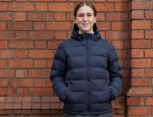 A girl stood against a brick wall wearing school uniform and a puffer coat by 1880 Club