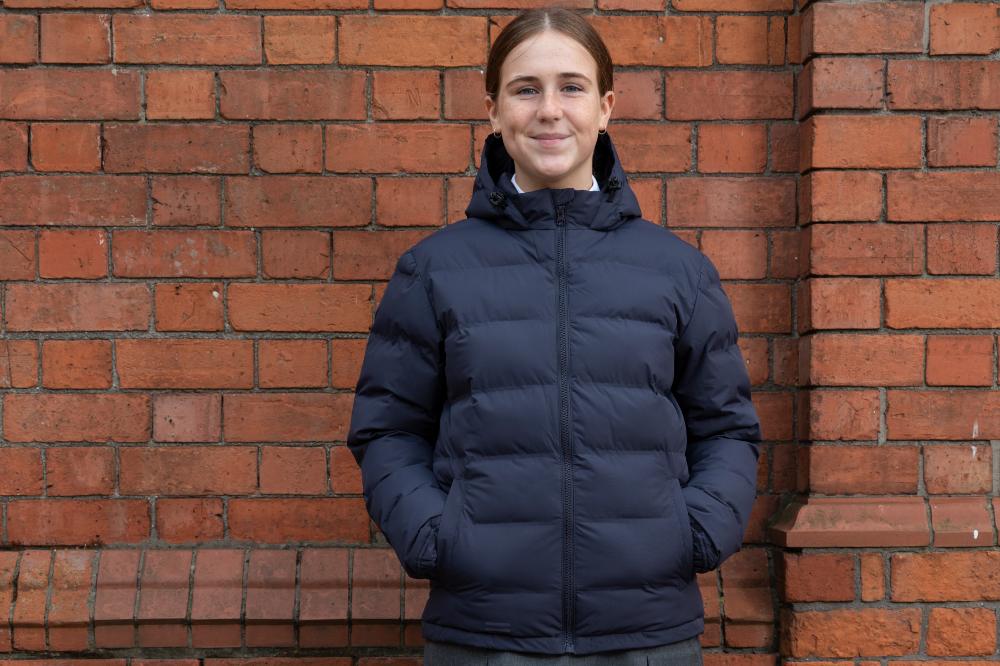 A girl stood against a brick wall wearing school uniform and a puffer coat by 1880 Club