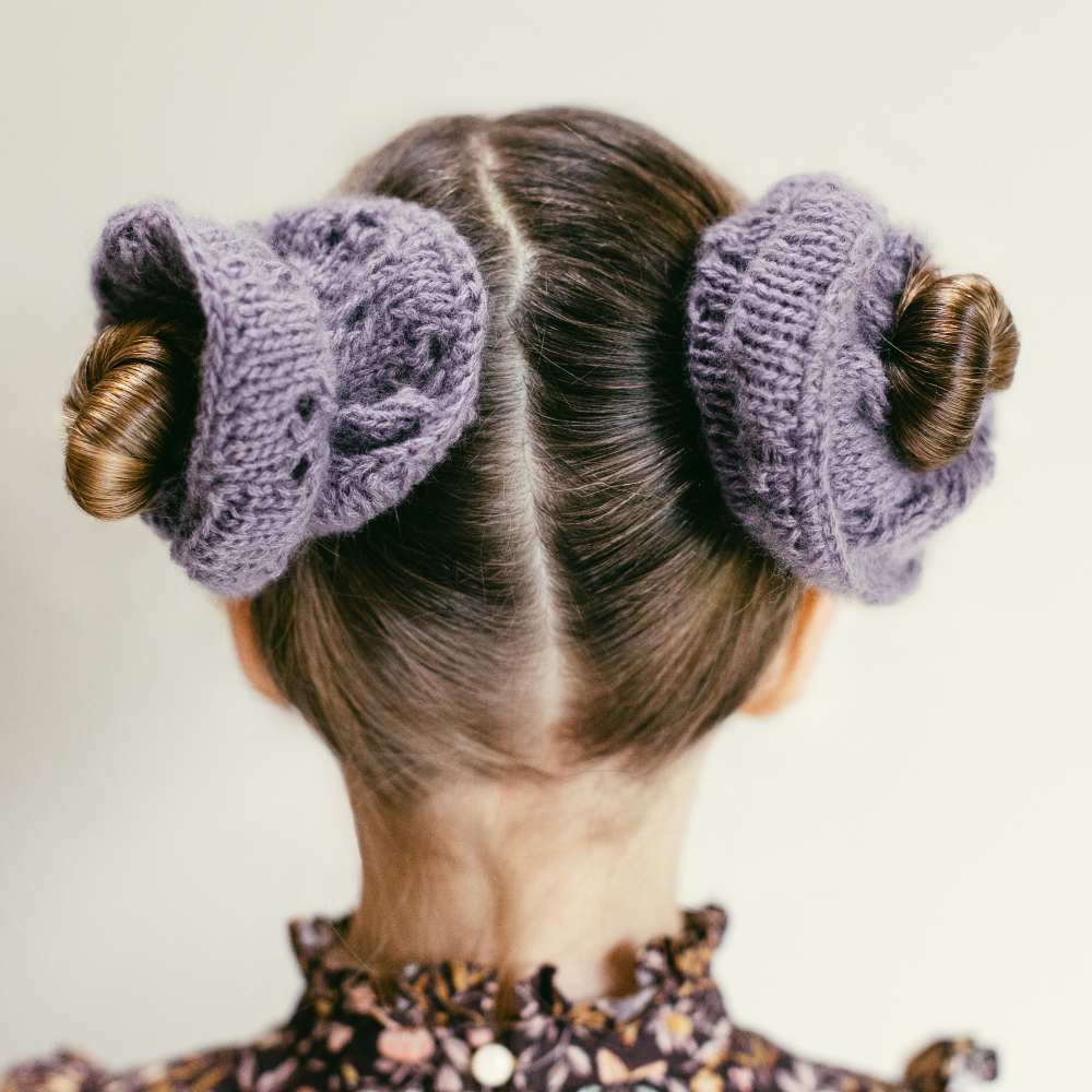 The back of a child's head with their hair in buns wrapped in lilac knitted scrunchies 